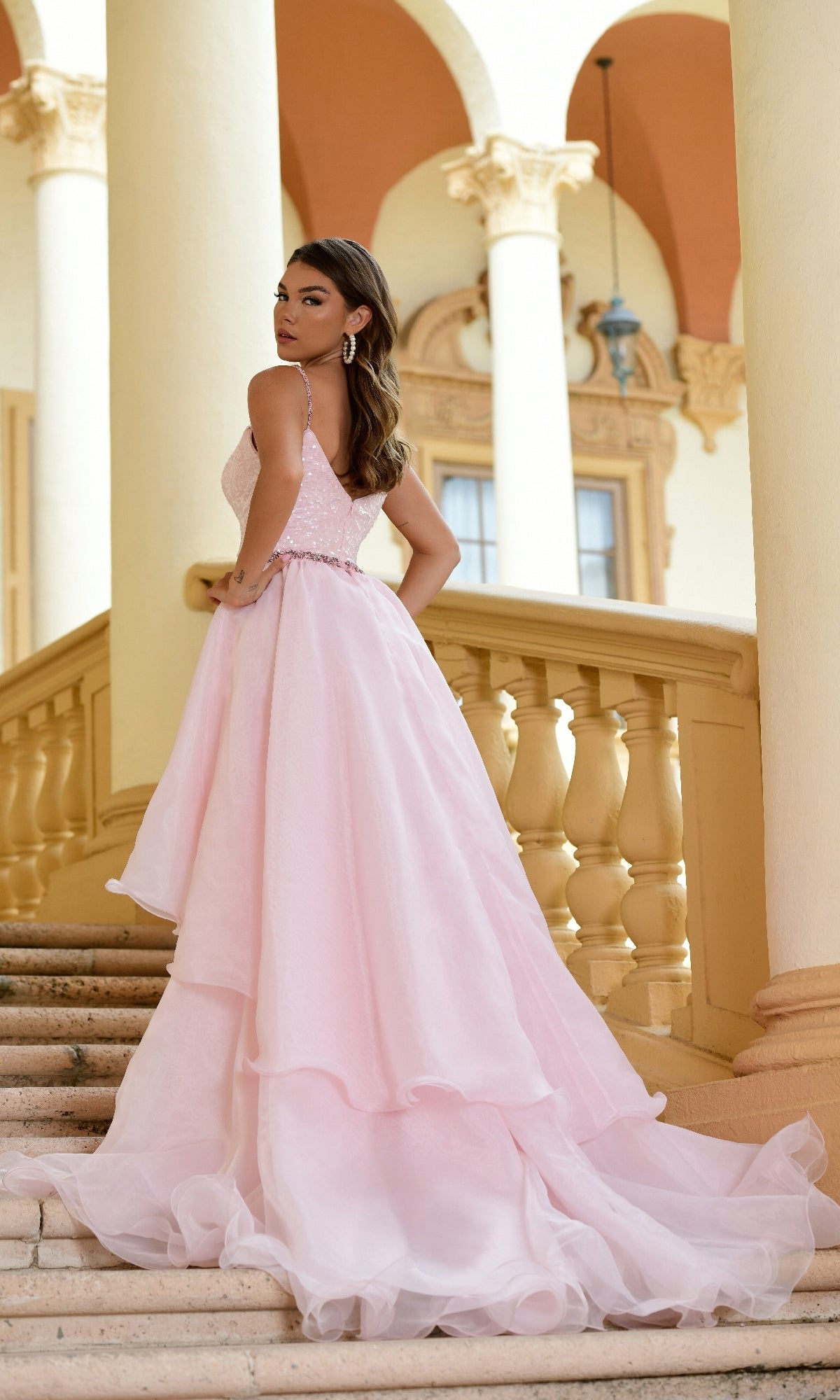 A woman in the Long Formal Dress 28560 by Ava Presley, a light pink ball gown with a chiffon overskirt, stands on stone steps before ornate columns and arches, glancing over her shoulder.